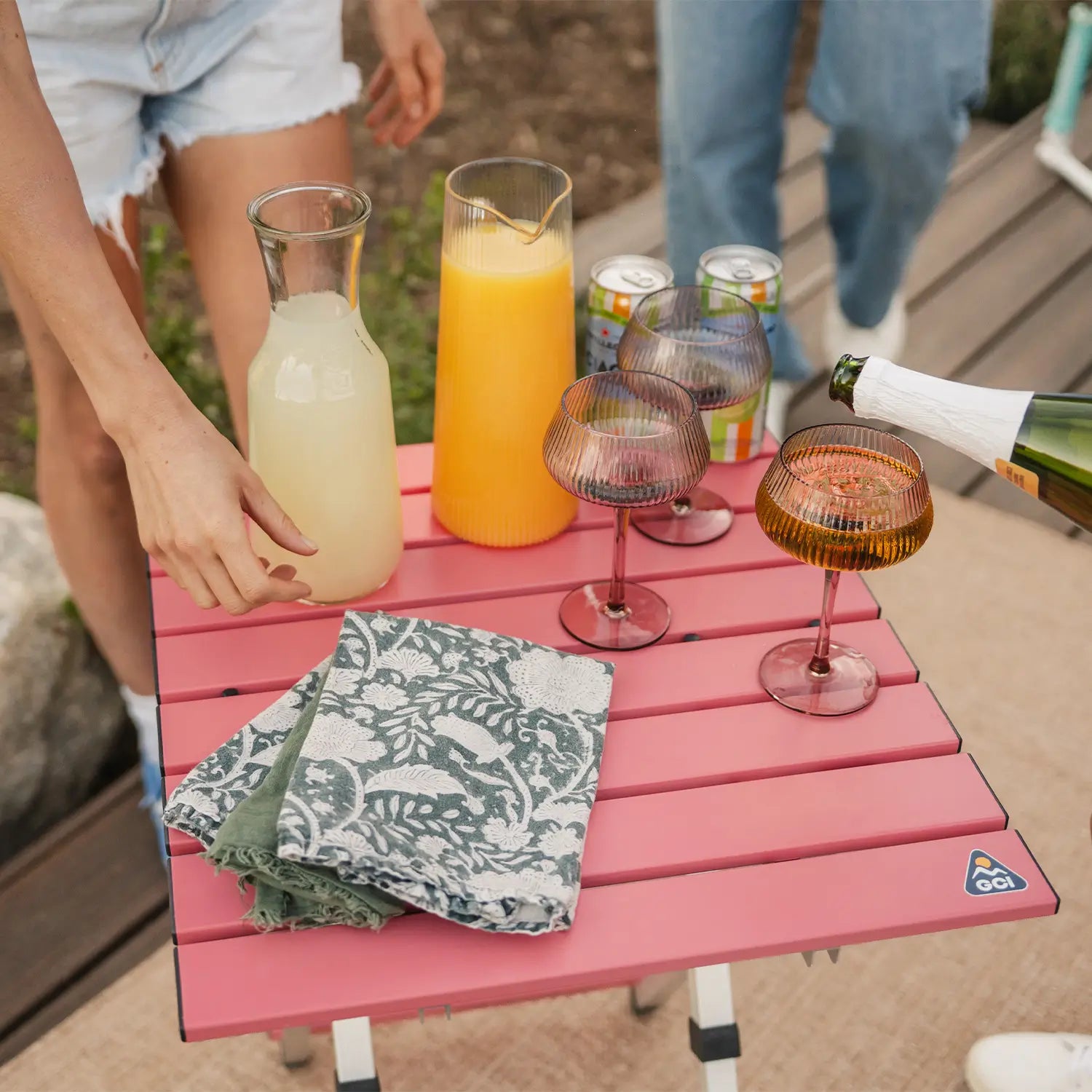 A woman pouring a glass of juice into a cup thats positioned on a coral Compact Camp Table. 