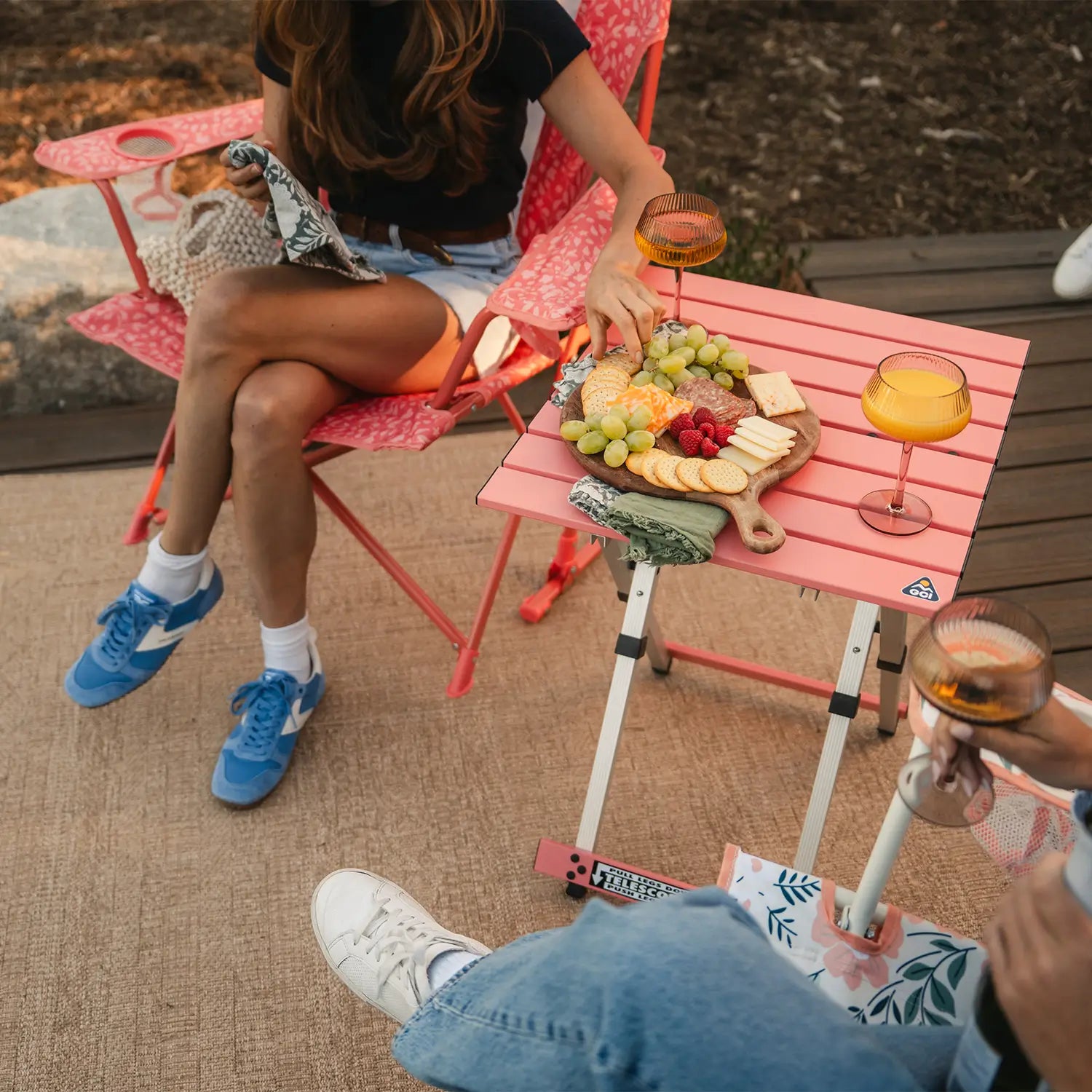 Two women sitting in coral Comfort Pro Rockers and having a snack thats positioned on a coral Compact Camp Table. 