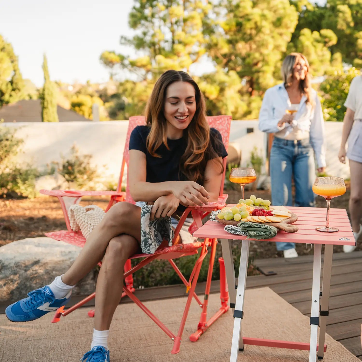 A woman sitting in a coral Comfort Pro Rockers is grabbing a snack resting on a coral Compact Camp Table. 