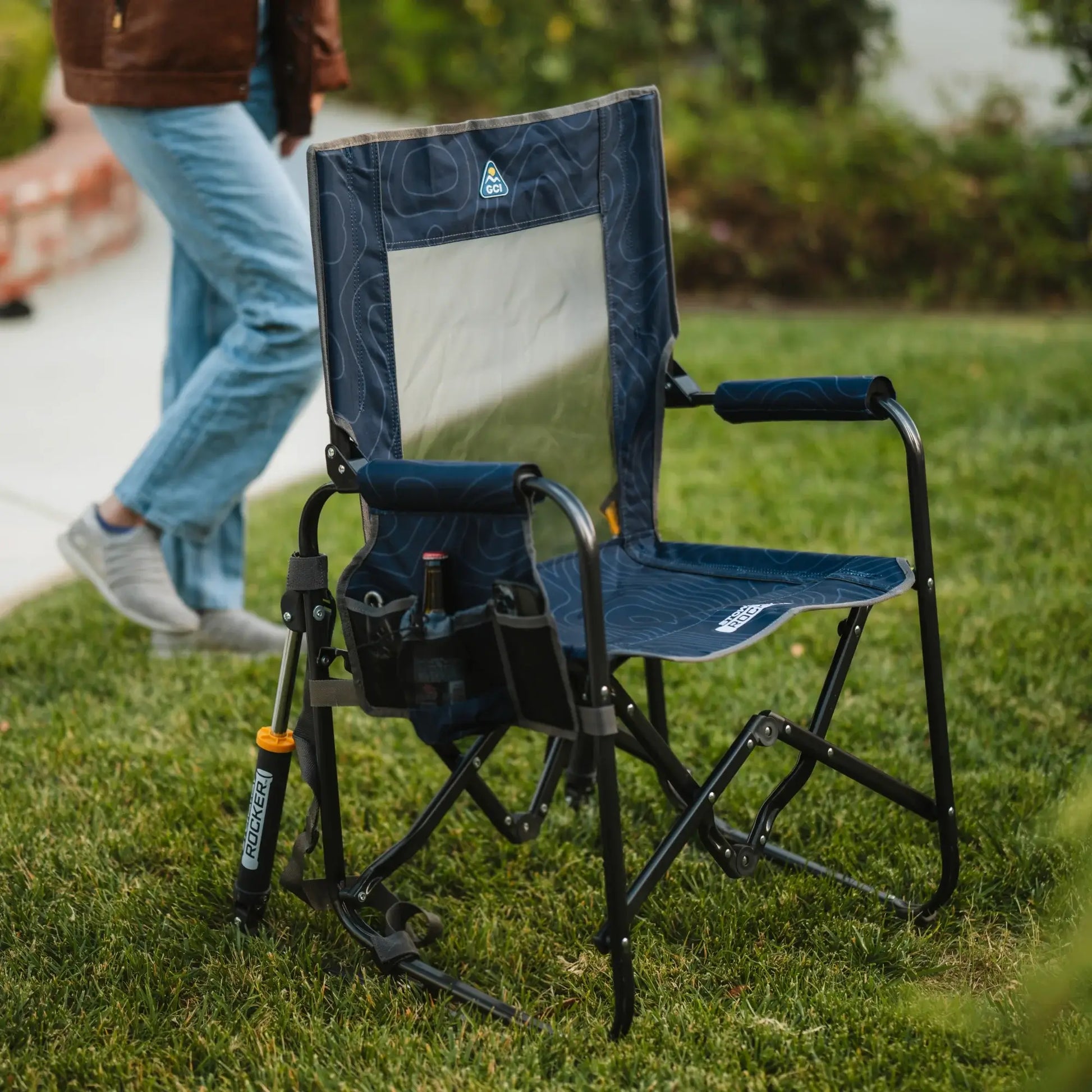 A navy topo Stowaway Rocker unfolded on a patch of grass while a person walks closely behind the chair. 