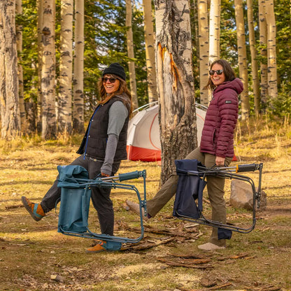 Two people carrying folded Teal Grab & Go Rockers through a wooded campsite.