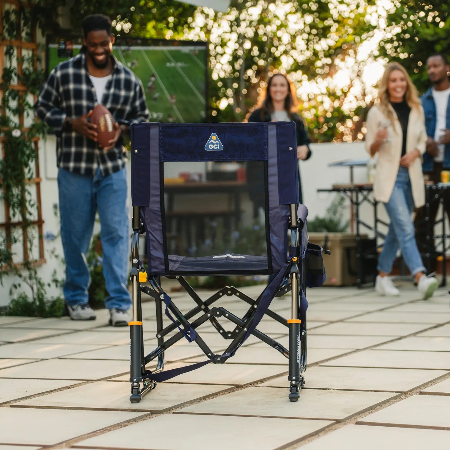 A tailgater rich blue Stowaway Rocker positioned in a backyard with people standing around watching television. 
