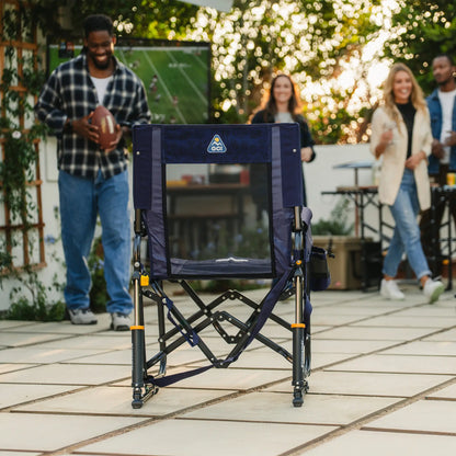 A tailgater rich blue Stowaway Rocker positioned in a backyard with people standing around watching television. 