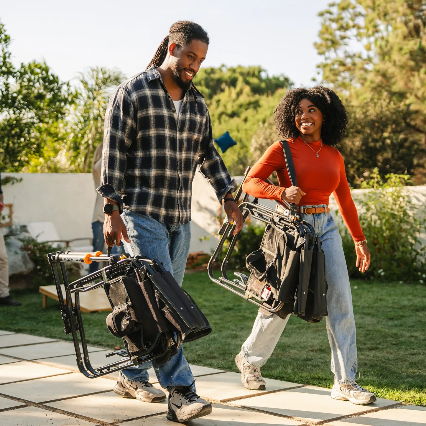 Two people carrying tailgater dark charcoal Stowaway Rockers in a backyard. 