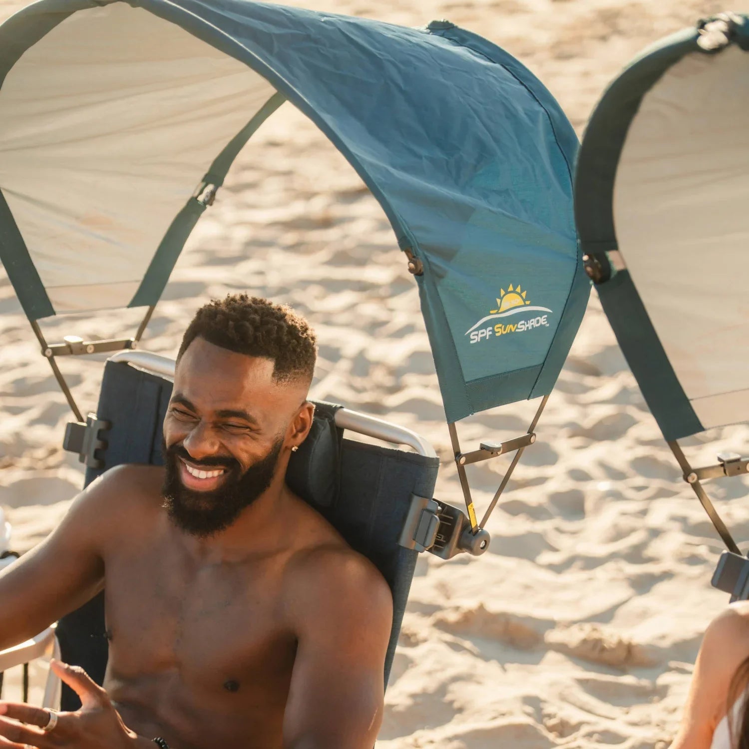 Man smiling beneath the arched canopy of a teal GCI Outdoor SPF SunShade Accessory clamped to his beach chair.